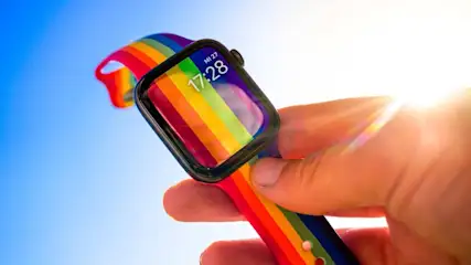 Eine Hand hält eine Smartwatch mit einem Regenbogenarmband vor einem blauen Himmel, die 17:28 anzeigt.