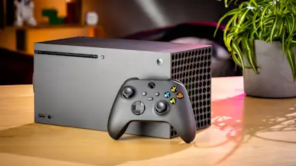 Ein grauer Xbox-Konsole mit Controller steht auf einem Holztisch, neben einer Topfpflanze.