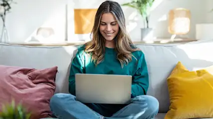 Eine junge Frau sitzt im Schneidersitz auf einem Sofa und blickt lächelnd auf ihren Laptop. Sie trägt einen petrolfarbenen Pullover und ist in einem sonnendurchfluteten Wohnzimmer.