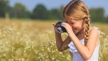 Ein Kind in weißem Top hält eine Kamera in einem Feld mit weißen Blumen und Bäumen unter blauem Himmel.