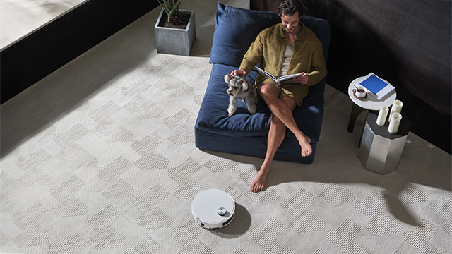  Mann sitzt entspannt auf einem blauen Sessel auf hell gemustertem Teppich, liest ein Buch und streichelt einen kleinen Hund, während im Vordergrund ein weißer Saugroboter selbstständig den Boden reinigt.