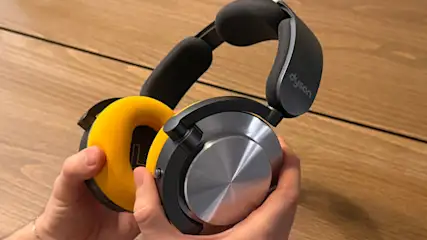 Eine Person hält Dyson Kopfhörer mit gelben Ohrpolstern und einem grauen Kopfbügel vor einem Holzhintergrund.