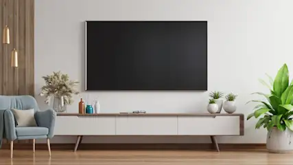Ein Wohnzimmer mit einem großen schwarzen Fernseher an einer weißen Wand über einem weißen und holzfarbenen Sideboard. Ein blauer Sessel ist links.