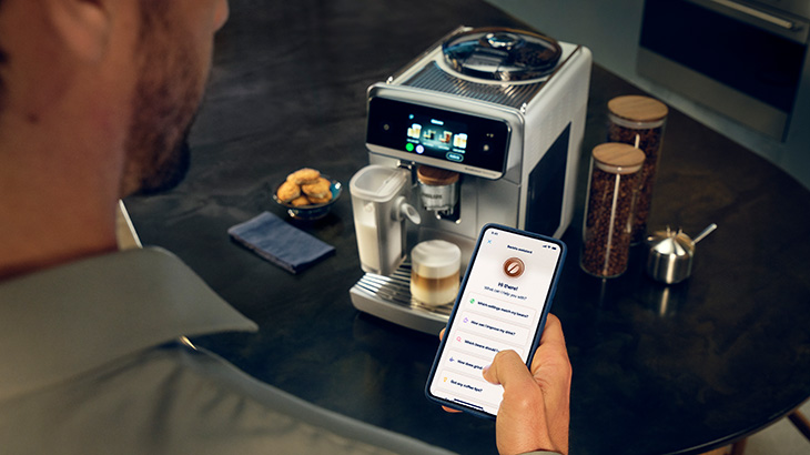 Eine Kaffeemaschine im Hintergrund, ein Mann der ein Handy mit geöffneter App hält im Vordergrund.