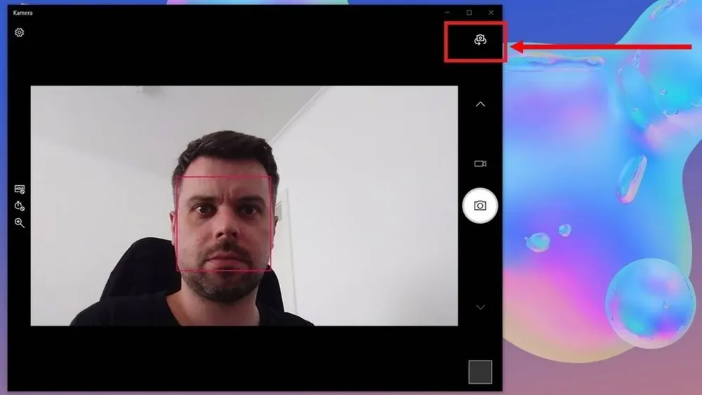 Ein Mann vor einer Kamera-App mit roter Gesichtserkennung, vor einem weißen Hintergrund. Die App hat eine Einstellungsoption. Rechts ein Pfeil auf das Symbol, um die Kamera umzukehren.