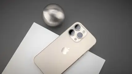 Ein goldenes Smartphone liegt auf Papier neben einer Metallkugel auf grauem Hintergrund. Das Telefon hat drei Kameras.