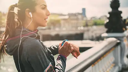 Frau in Sportkleidung mit Kopfhörern blickt auf eine blaue Smartwatch vor einer Stadtkulisse und einem Geländer.