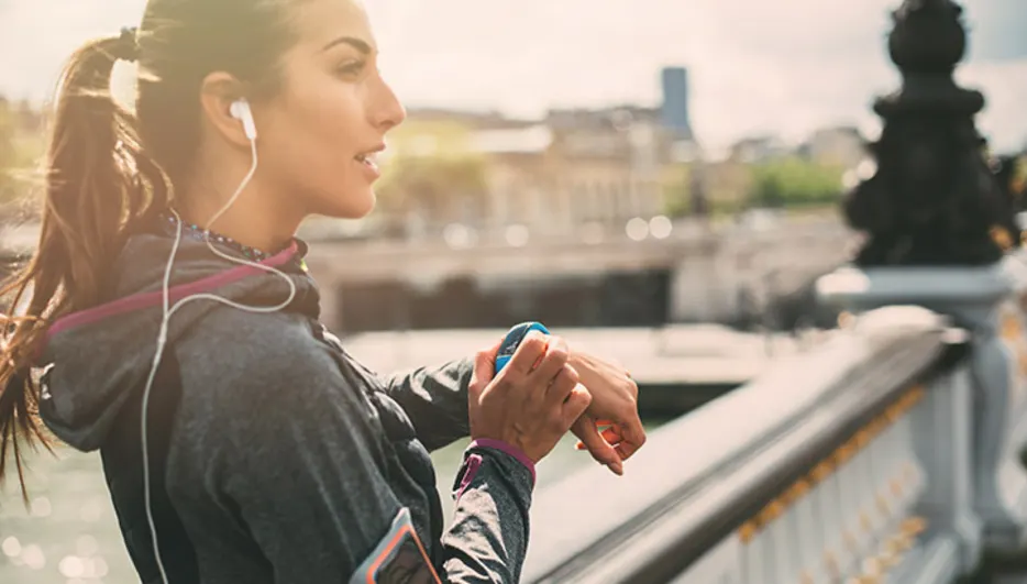 Frau in Sportkleidung mit Kopfhörern blickt auf eine blaue Smartwatch vor einer Stadtkulisse und einem Geländer.