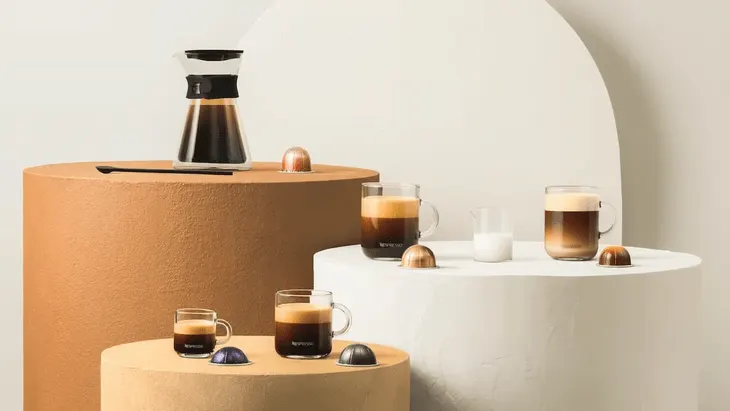 Auf Staffeleien stehen Kaffeekaraffe, Tassen mit Kaffee und Milch, Kapseln und ein Krug mit Milch vor hellem Hintergrund.