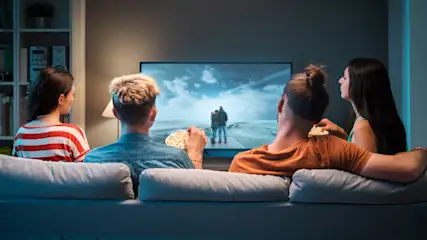 Vier Personen sitzen auf einem grauen Sofa und schauen auf einen großen Fernseher. Zwei Personen essen Popcorn.