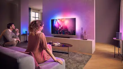 Wohnzimmer mit Paar auf lila Couch, das einen Konzertfilm auf einem beleuchteten Fernseher ansieht. Tisch mit Büchern und Flaschen.