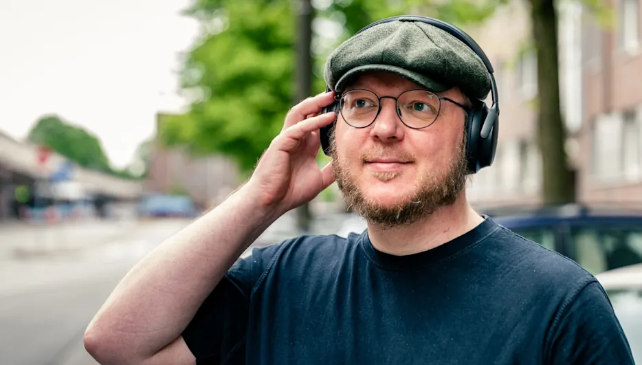 Andreas trägt den Bose QuietComfort Ultra an einer Bushaltestelle und blickt auf sein Smartphone.