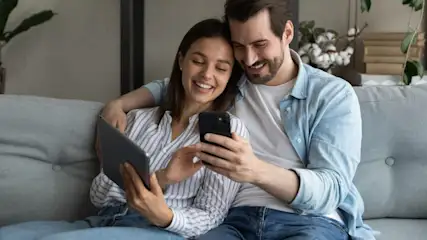 Ein Paar sitzt auf einer grauen Couch und betrachtet ein Tablet und ein Smartphone.