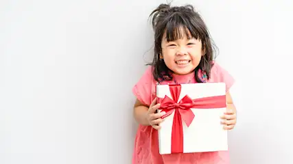 Ein lächelndes Kind im rosa Hemd hält eine weiße Geschenkbox mit einer roten Schleife vor einem weißen Hintergrund.