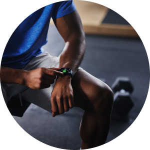 Person im Fitnessstudio, die auf einer Bank sitzt und ihre Sport-Smartwatch am Handgelenk bedient, im Hintergrund liegen Hanteln auf dem Boden.