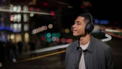 Junger Mann mit Over-Ear-Kopfhörern Sony WH-1000XM5 genießt Musik mit geschlossenen Augen. Im Hintergrund verschwommene Lichter einer Stadt.