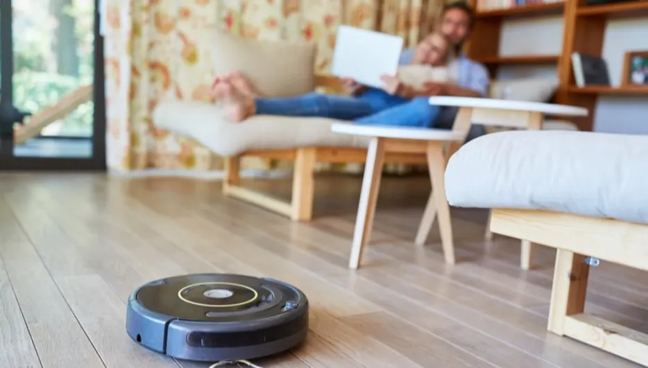 Ein Roboterstaubsauger reinigt einen Holzboden. Im Hintergrund sitzen zwei Personen auf einer Ottomane und lesen.
