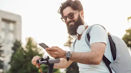 Ein Mann mit Bart und Sonnenbrille benutzt ein Handy auf einem Roller. Er trägt Kopfhörer und einen Rucksack. Gebäude und Bäume im Hintergrund.