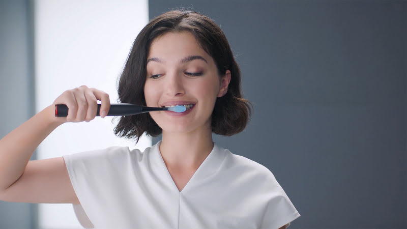 Eine Frau in Weiß putzt Zähne mit einer schwarzen elektrischen Zahnbürste vor einem grauen Hintergrund.