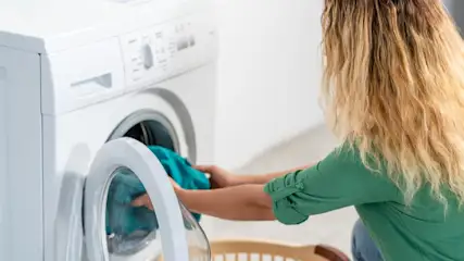 Eine Person in einem grünen Oberteil leert blaue Wäsche aus einer weißen Waschmaschine in einen Wäschekorb.
