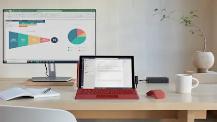 Ein Schreibtisch mit einem roten Laptop, einem Monitor mit Diagrammen, einer weißen Tasse, einer Maus und einer Pflanze.