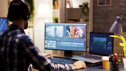Mann mit Kopfhörern vor Computern mit Videoschnittsoftware. Auf dem Schreibtisch stehen Stifte und ein Kaffeebecher.