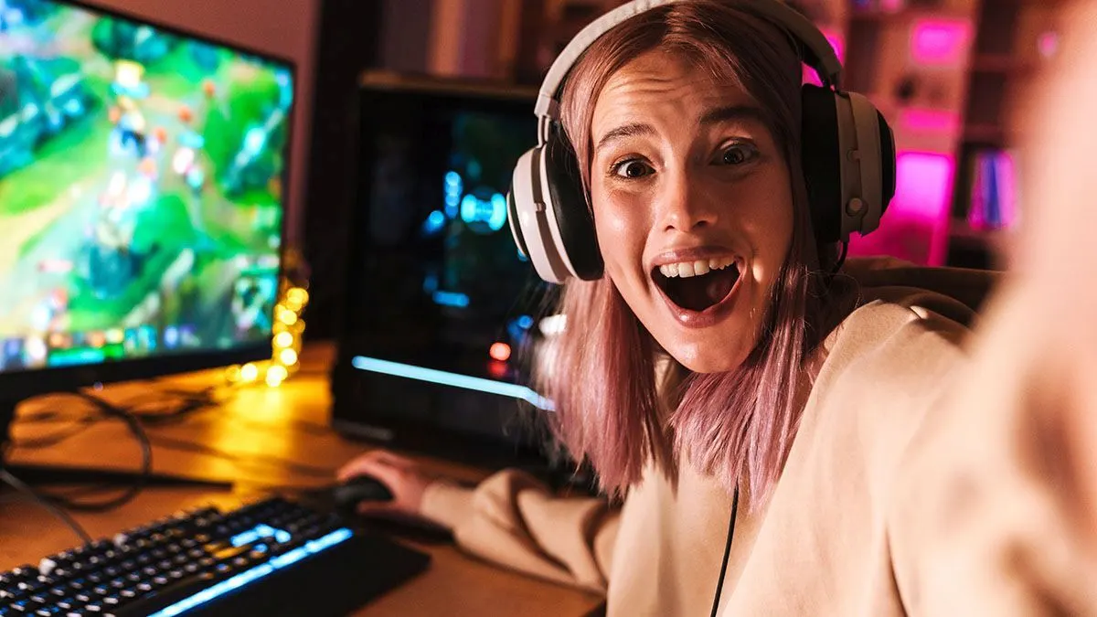 Eine junge Frau sitzt vor einem Gaming-PC und schaut begeistert zum Betrachter, auf dem Monitor ist ein Spiel zu sehen.