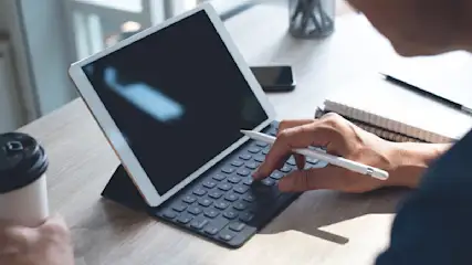 Eine Person tippt mit einem Stift auf die Tastatur eines Tablets. Ein Notizbuch, ein Stift, ein Telefon und ein Kaffee stehen auf dem Tisch.