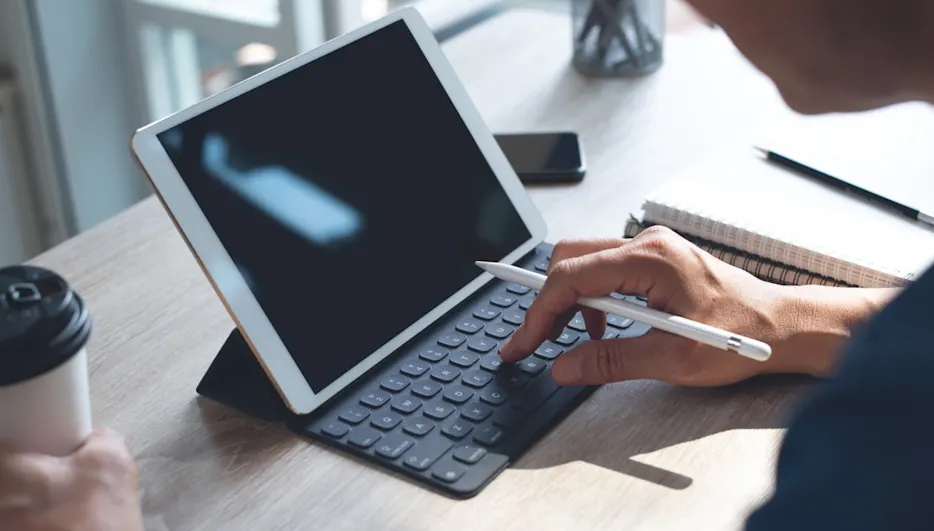 Eine Person tippt mit einem Stift auf die Tastatur eines Tablets. Ein Notizbuch, ein Stift, ein Telefon und ein Kaffee stehen auf dem Tisch.