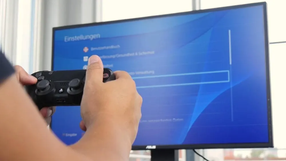 Eine Person hält einen schwarzen PlayStation-Controller vor einem Monitor mit blauem Einstellungsmenü.