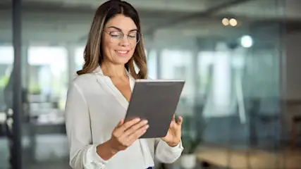 Eine Frau mit Brille und weißer Bluse hält ein graues Tablet in einem Büro mit Glaswänden.