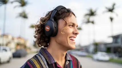 Ein Mann mit Kopfhörern und gestreiftem Hemd lächelt. Autos und Palmen sind im Hintergrund verschwommen dargestellt.