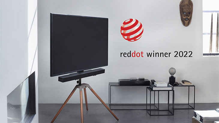Ein Fernseher steht auf einem Dreibeinstativ vor einer Wand mit Dekoration und dem Text "reddot winner 2022". Tische stehen daneben.