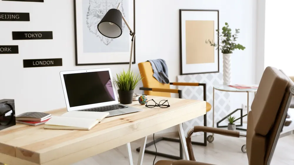Ein Schreibtisch mit Laptop, Notizbüchern und einer Brille, beleuchtet von einer Lampe. Im Hintergrund ein Stuhl und Dekoration.
