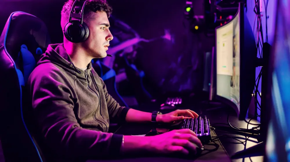 Ein Mann mit Kopfhörern sitzt an einem Computer mit Tastatur und Maus vor einem Monitor. Blaue und violette Beleuchtung im Hintergrund.