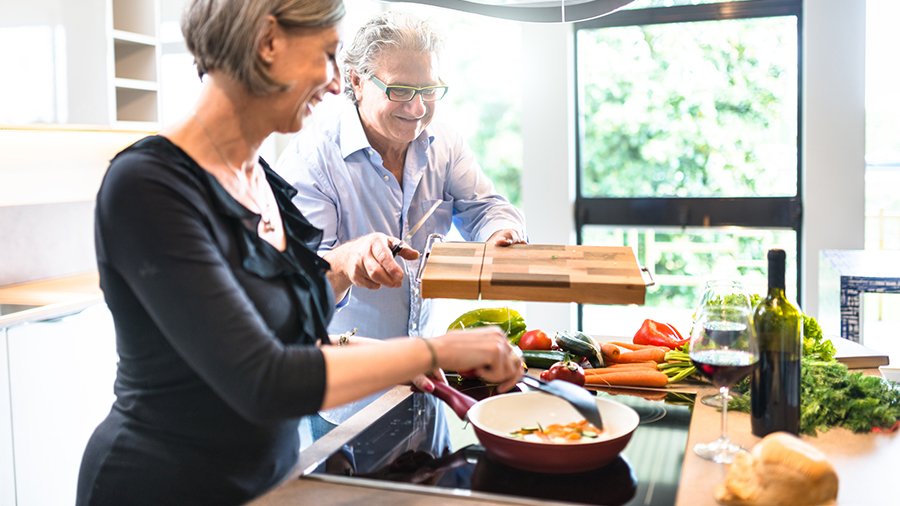 In der Küche kocht eine Frau mit einem Mann. Gemüse, Wein und ein Schneidebrett stehen auf der Arbeitsfläche.