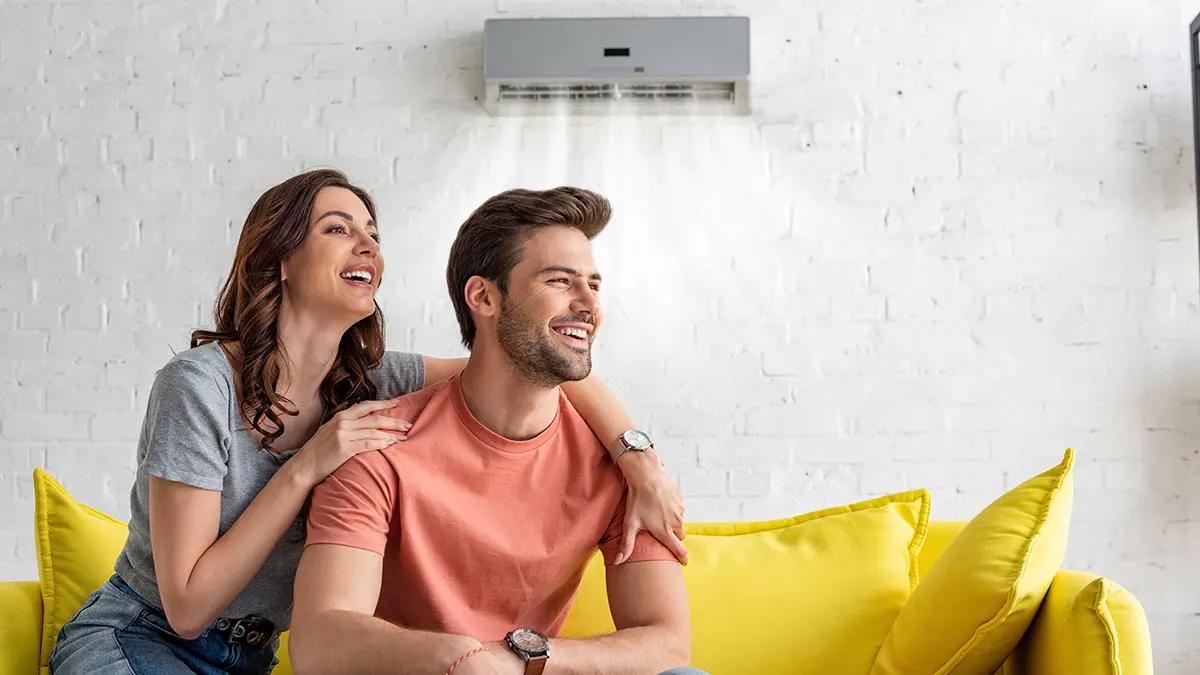 Junges Paar sitzt lachend auf gelbem Sofa. Eine Klimaanlage ist an der weißen Backsteinwand im Hintergrund angebracht.