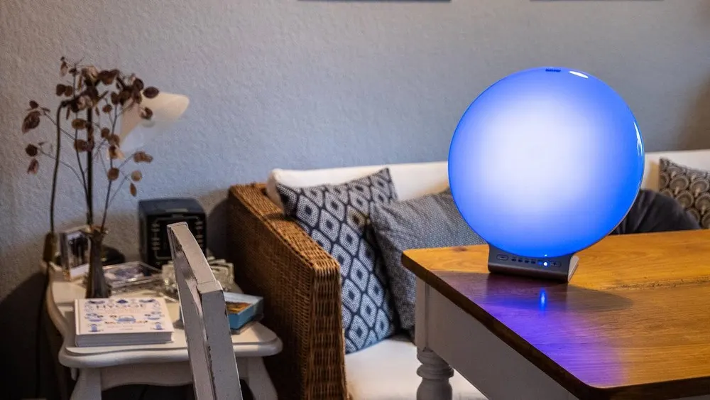 Blaue Tageslichtlampe auf einem Holztisch, Sofa mit Kissen im Hintergrund. Ein Beistelltisch mit Deko und Buch ist auch sichtbar.