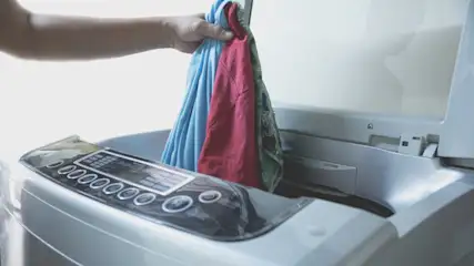 Eine Hand legt blaue und rote Wäsche in eine weiße Waschmaschine mit digitaler Steuerung.