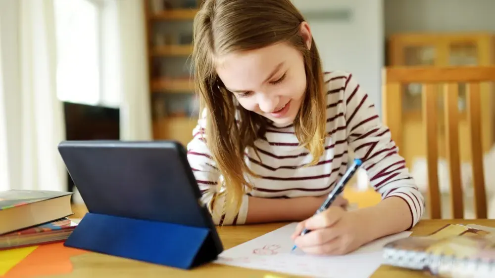 Ein Mädchen erledigt seine Hausaufgaben mit Stift, Papier und Tablet an einem Tisch.