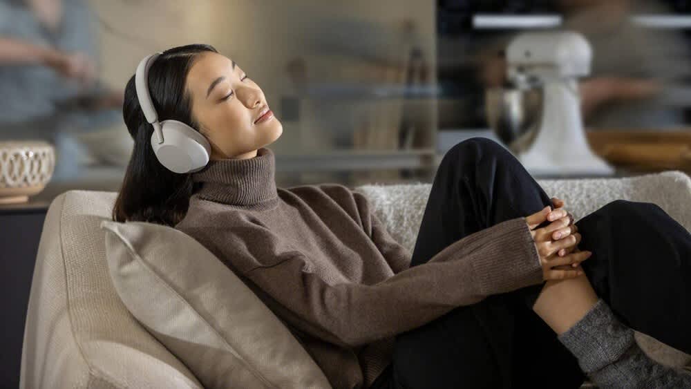 Frau liegt mit Over-Ear-Kopfhörern Sony WH-1000XM5 auf der Couch und hört Musik