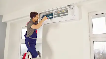 Ein Mann in blauer Arbeitskleidung steht auf einer Leiter und repariert eine weiße Klimaanlage an einer Wand.