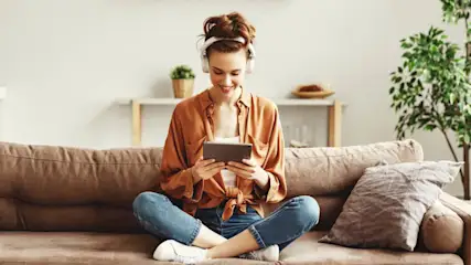 Frau mit Kopfhörern sitzt im Schneidersitz auf einem braunen Sofa und hält ein Tablet in den Händen. Im Hintergrund sind Regale und Pflanzen.