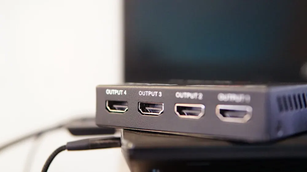 Nahaufnahme eines schwarzen HDMI-Splitters mit vier Ausgängen, beschriftet mit "OUTPUT 4" bis "OUTPUT 1", auf einem dunklen Untergrund. Ein Kabel ist angeschlossen.