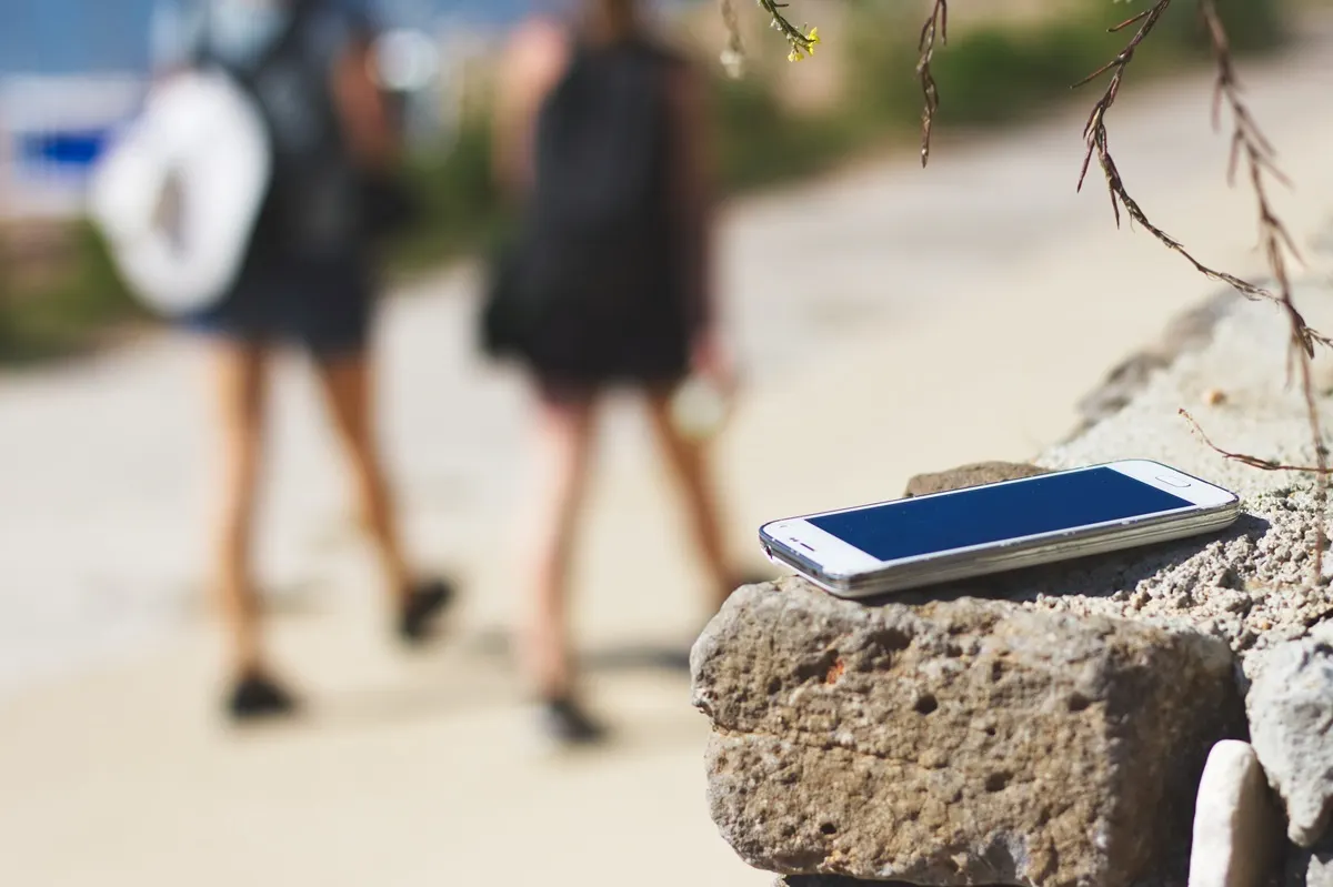 Ein blaues Smartphone liegt auf einem Stein, im Hintergrund gehen zwei Personen in dunkler Kleidung entlang eines Weges.