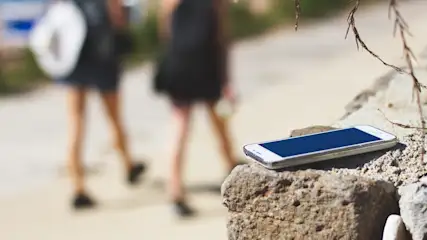 Ein blaues Smartphone liegt auf einem Stein, im Hintergrund gehen zwei Personen in dunkler Kleidung entlang eines Weges.