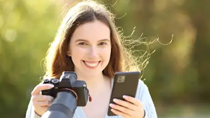 Eine Frau hält eine Kamera und ein Smartphone vor einem grünen Hintergrund.