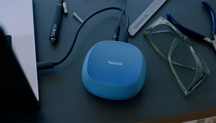 Blaues SanDisk-Gerät mit zwei Kabeln auf dem Schreibtisch; daneben Lineal, Schraubendreher, Bit und Schutzbrille.