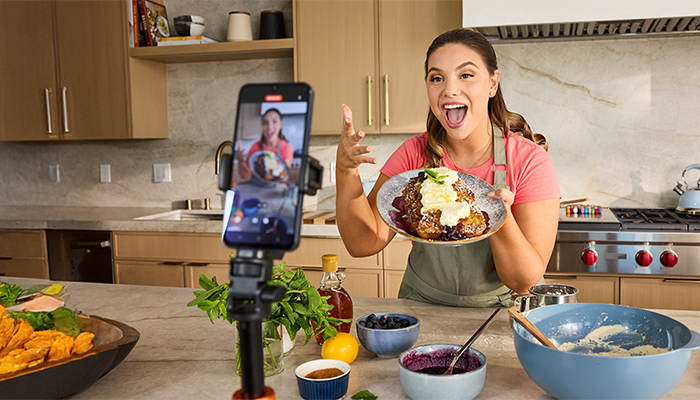 Person präsentiert Pancakes mit Beeren und Sahne in der Küche; Smartphone auf Stativ filmt, Schüsseln und Zutaten auf der Arbeitsplatte.