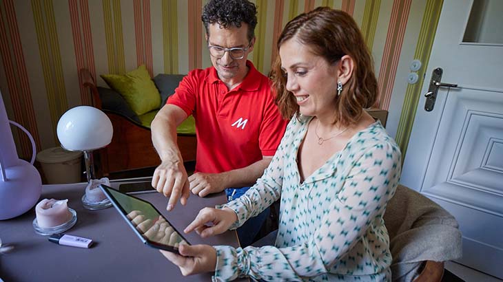 Eine Frau und ein Mann, beide an einem Tisch, bedienen ein Tablet. Der Mann trägt ein rotes Poloshirt, die Frau eine Bluse mit Muster.